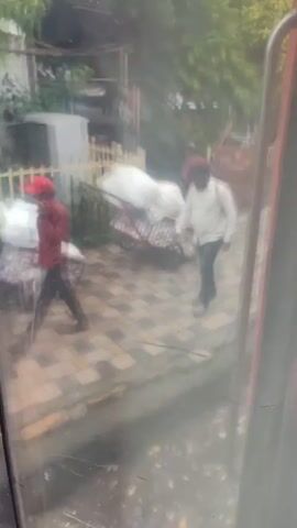 A brief video clip captures two individuals pushing carts loaded with large white bundles. The scene appears to be a train platform, with a red train carriage visible in the foreground. The platform is paved with interlocking tiles, and a metal fence separates it from lush greenery. Another person is seen walking on the platform in the background, and a third person is visible further down the platform. The overall ambiance suggests an outdoor, possibly busy, location.
