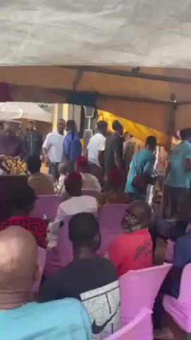The video depicts a gathering of people outdoors, likely under a tent or canopy. Many are seated on pink plastic chairs, while others stand or move around. People of various ages are present, including adults and children. Some are wearing traditional attire, such as red caps. A man in a purple outfit stands near the front, seemingly addressing the crowd or observing the event. Behind him, a girl in a yellow top and pink skirt is visible. The environment appears to be a yard or open space, with greenery and other structures in the background. A speaker can be heard playing music. The camera angle shifts and blurs, but generally captures a wide view of the event and the people present.