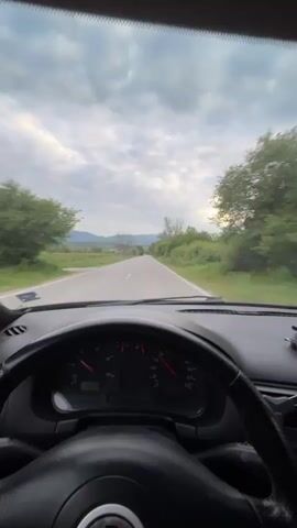 The video shows a person driving on an open road. The view is from the driver's perspective, showing the steering wheel, dashboard, and the road ahead through the windshield. The car is traveling on a paved road with fields and sparse trees on either side. In the distance, there are rolling hills. The sky is overcast. The driver's hand is visible on the steering wheel. The car's speedometer is partially visible. The camera pans slightly to the left at one point, showing the side mirror.