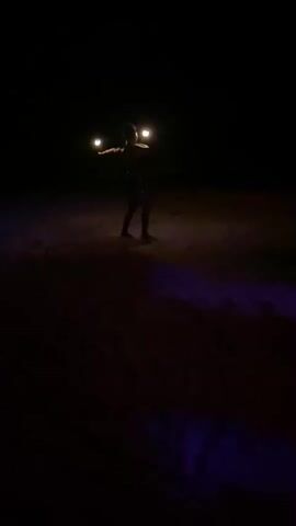 A person is performing a fire dance on a sandy beach at night. The performer, who appears to be a woman, is wearing a dark top and bottoms and is holding two flaming objects, presumably fire torches. The beach is dark, but the fire and possibly some ground lighting illuminate the immediate area around the performer, casting a warm glow on the sand. The scene has a dynamic feel due to the spinning and movement of the fire torches. The overall impression is of an outdoor performance in a natural setting.