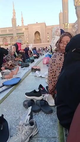 The video shows a large group of women sitting on mats on the marble ground outside a large building with tall columns and minarets. The women are dressed in various styles of modest clothing, including headscarves and long dresses. Many of them have their shoes removed and placed in front of them. Some of the women are reading books or looking at their phones, while others are talking to each other. There is a baby lying on a mat in the foreground. The overall atmosphere appears peaceful and serene.
