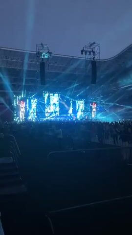 A wide shot captures a concert stage in a stadium at dusk. The stage is illuminated with bright blue lights and features large screens displaying abstract visuals. Two figures, presumably performers, are visible on the stage, each standing on a raised platform and facing the audience. A large crowd of spectators is gathered in front of the stage, many holding up their phones. The stadium seating is visible in the background, and the sky above is a dim, twilight blue. There are metal barriers and some security personnel scattered throughout the audience area.