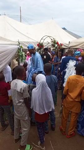 Here is a concise description of the video:

The video shows a group of children, likely wedding guests, watching a wedding ceremony under a white tent. An officiant speaks while holding a phone. Many people, including women in colorful headscarves, can be seen in the background. The wedding arch is decorated with flowers.