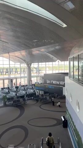 Here is a description of the video:

The video shows the inside of an airport terminal. The airport is bright and modern, with large windows and a high, perforated ceiling. There are rows of empty seats in the waiting area. Several people are walking around, some with luggage. Signage directs passengers to different destinations. Outside the windows, the airfield and a plane are visible.