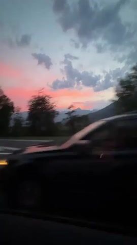 The video captures a scenic view from a moving vehicle, likely a car, looking out the window at a sunset over a mountainous landscape. The sky is a vibrant mix of pink, orange, and blue hues, with scattered clouds adding texture. In the foreground, a highway with white lane markings is visible, indicating the vehicle's forward motion. Trees and distant power lines dot the landscape, suggesting a semi-rural or industrial area. The overall impression is one of a journey, with the natural beauty of the sunset providing a dramatic backdrop.