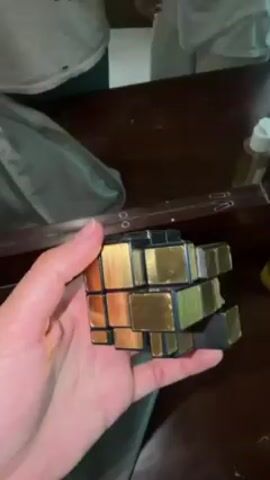 A hand holds a gold and black Mirror Cube in front of a mirror. The cube is not in a solved state, as the cuboids are not aligned. In the reflection in the mirror, two people are seen. One person is wearing a white shirt and is using a white cloth to wipe a dark-colored surface, presumably a table. The other person is partially visible in the reflection. A bottle is also visible on the table. The cube is turned in the hand, revealing different sides.