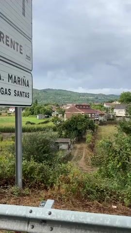 The video shows a rural landscape under a cloudy sky. In the foreground, a metal railing is visible. Behind the railing, there are green bushes and trees, with some bare branches on a tree in the middle ground. Further back, there are fields, agricultural plots, and scattered buildings that form a small village. In the distance, a hazy mountain range is visible, with clouds obscuring its peak. The overall impression is one of a slightly overcast day in a country setting. There are no people or discernible actions in the video.
