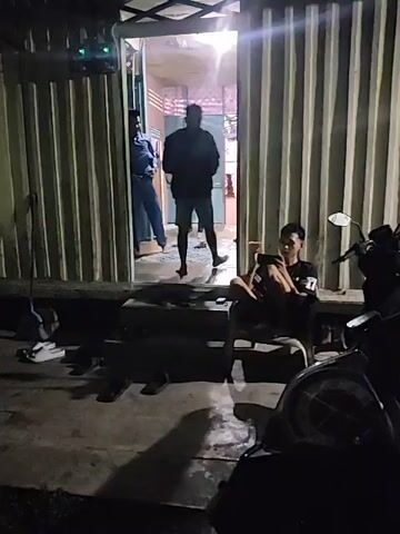 The video captures a dimly lit exterior scene at night. In the foreground, a person is seated on a chair to the right, holding a phone and appearing to smoke. A motorcycle is parked to the right of the frame, partially obscuring the view of the seated individual. In the background, a corrugated metal structure with an open doorway is visible. Three individuals are present near the doorway. One person, wearing a blue shirt, stands to the left inside the doorway. Another person, wearing dark clothing, stands further inside the doorway. A third person, also in dark clothing, stands slightly further into the structure. The floor area near the doorway appears to have some debris. Steps lead up to the doorway, with several pairs of sandals placed on them. A broom is leaning against the corrugated wall to the left of the doorway. The overall environment suggests a casual, possibly outdoor gathering or waiting area at dusk or night.