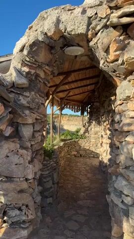 The video begins with a view through a rustic stone archway. The path ahead is paved with stones and flanked by rough stone walls. Sunlight streams through the arch, illuminating the scene. As the camera moves forward, it enters a covered walkway with a wooden roof supported by rough-hewn wooden beams. The walkway overlooks a natural landscape featuring green hills and a quarry. Lush green foliage grows along the stone walls on the left side of the walkway. The sky is a clear blue. The camera continues to move through the walkway, offering a panoramic view of the surrounding environment.