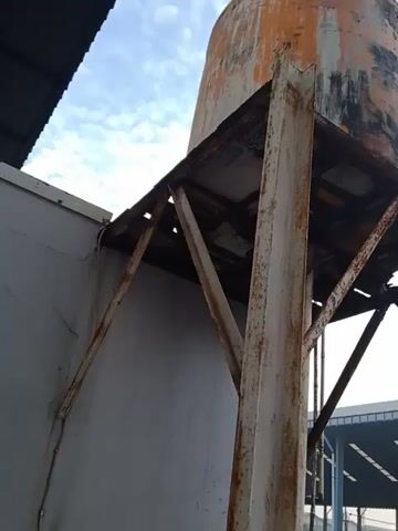 The camera is pointed upwards towards a water tank mounted on a rusty metal frame against a cloudy sky. A building with a metallic roof is visible in the background. The tank is cylindrical, appears old, and has peeling paint. The supporting frame is composed of intersecting metal beams, displaying significant rust. There are also blue pipes and a valve within the frame, connected to the tank. The camera's perspective is from below, looking up at the structure.