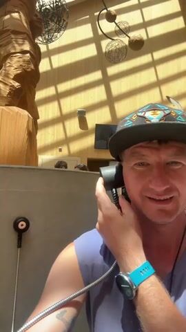 This is a selfie-style video of a man holding a telephone receiver to his ear, looking at the camera with a smile. He is wearing a dark baseball cap with a colorful, possibly Native American-inspired, pattern, and a grey t-shirt. A blue fitness tracker is visible on his left wrist. Behind him, a large, intricately carved wooden sculpture of a human figure is prominently displayed. The sculpture is illuminated by natural light filtering through a sloped, wooden-beamed ceiling with skylights. Decorative spherical objects hang from the ceiling. In the background, two people can be seen peeking over a counter. There's a grey wall with a metal fixture that has a black circular object and a coiled silver cord, suggesting it's part of an older telephone system. A small, informational plaque is affixed to the wall to the left of the sculpture. The overall setting appears to be a public indoor space, possibly a museum or a lodge, given the artwork and architecture. The lighting suggests it is daytime.