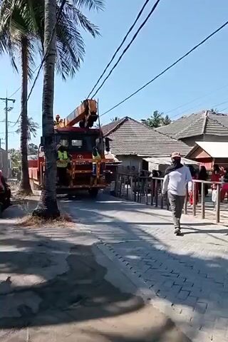 A red truck with a crane is parked on a paved road, facing away from the camera. Several individuals wearing yellow safety vests are visible on and around the truck. The crane is extended upwards, appearing to be in use. A person in a white long-sleeved shirt and dark pants is walking away from the truck on the right side of the road. The environment appears to be a street with buildings and palm trees. The sky is clear and blue.