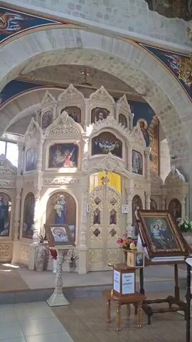The video opens with a view of an ornate altar and a wall of icons in what appears to be a church. The camera pans up to reveal a large, unfinished ceiling with exposed stone and a partially completed mural. A chandelier hangs from the ceiling. The video then pans back down to the altar, focusing on the details of the icons and the altar itself. The scene is well-lit, and the overall impression is one of religious devotion and historical significance.