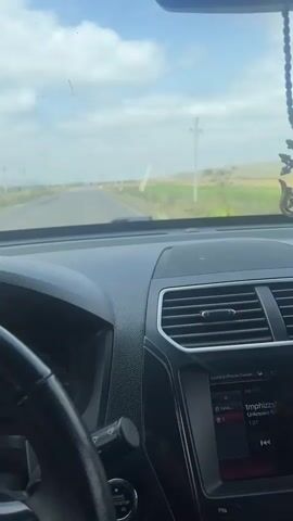 The video shows the interior of a car as it drives down a road. The perspective is from the driver's seat. The dashboard is visible, with the steering wheel on the left. A person's hand is on the steering wheel. The radio is on and displays the song "tmphizzh". Outside the car, the road is flanked by green fields and utility poles. The sky is cloudy with some blue patches. There is a beaded string of what appears to be prayer beads hanging from the rearview mirror. The car is moving forward on a paved road.