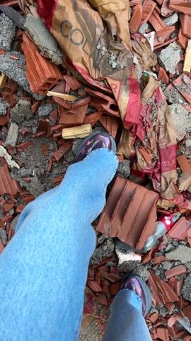 A first-person perspective video shows someone walking through a debris field. The person is wearing light blue jeans and colorful sneakers. The debris appears to be mostly broken red terracotta tiles and pieces of wood. A partially visible bag marked with "COL" is also present. The person steps over the debris, and their feet and legs are visible throughout the video. The environment looks like a construction or demolition site.