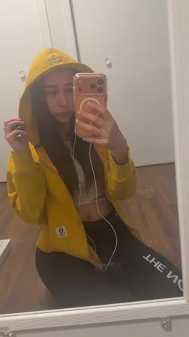 A young woman is kneeling in front of a mirror, taking a selfie. She is wearing a yellow zip-up hoodie with the hood up, a white crop top, and black leggings with white lettering. She is holding an iPhone in her right hand, which has a clear case and a MagSafe attachment on the back. White earphones are connected to the phone and dangling down. A wedding ring is visible on her left ring finger. The background consists of a white door with silver hardware, a wooden floor, and the mirror frame.