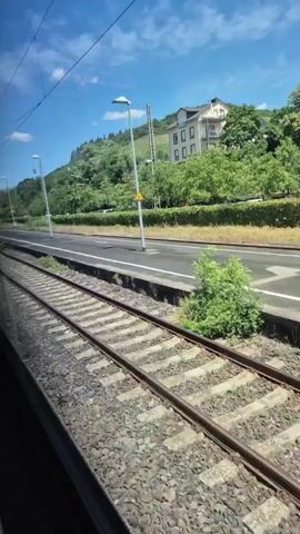 A train moves past a train station. The train tracks are in the foreground and the train station platform is visible in the midground. A building is visible in the background atop a hill covered in trees. The sky is blue with some clouds. There are streetlights and a utility pole along the platform. The camera is looking out of a train window.