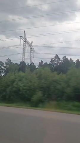This media file captures a view from a moving vehicle, likely on a road, with a forest bordering the right side of the frame. The sky is overcast with a layer of clouds, suggesting a dull or rainy day. A tall, metallic power line tower stands prominently in the background, with multiple electrical wires stretching across the sky. The forest is dense with a variety of trees, mostly green, indicating it is likely summer or late spring. A part of the power line appears to be damaged or in disrepair, with a long, white piece of material hanging down from it, possibly a torn section of insulation or a banner. The motion blur effect indicates the video was recorded while in transit. There is no visible text in the media file. The scene suggests a rural or semi-rural setting on the outskirts of Yekaterinburg, Russia.
