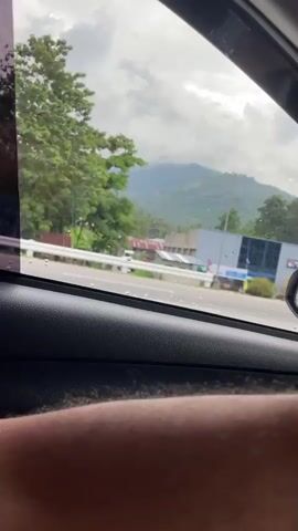 From inside a moving vehicle, a view from the passenger side window shows a highway. Outside, a mountain range with peaks obscured by clouds can be seen in the distance. Along the highway, there is a mix of commercial buildings, houses, and lush green vegetation, including trees and bushes. A white and yellow barrier separates the road from the landscaping. The car's side mirror is visible. A person's forearm is resting on the car's interior door panel. The overall tone is calm and scenic.