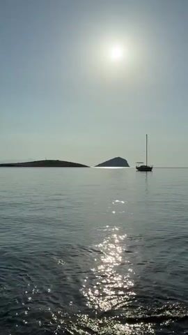 The video displays a tranquil seascape with a bright sun high in a clear sky. The sun's reflection shimmers on the calm water, creating a dazzling pathway. In the distance, two small islands are visible. The island on the left appears larger and flatter, while the one on the right is smaller and more conical. A solitary sailboat with its mast raised floats peacefully on the water between the viewer and the islands. The foreground shows the edge of the water, with gentle waves lapping against what appears to be a rocky shore. The overall impression is one of serene, sunlit calm.