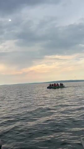 The video shows a raft with several people paddling on a large body of water. The sky is cloudy with the sun setting in the distance. The raft appears to be made of two inflatable pontoons connected by a frame. The people on the raft are wearing life jackets and are using paddles to propel themselves through the water. The scene is peaceful and serene, with the gentle rocking of the raft and the sound of the paddles breaking the surface of the water.