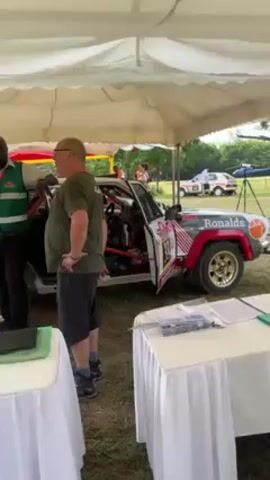 The video shows two men standing next to a rally car under a white tent. The man on the left is Black and wearing a green vest, black pants, and a badge. The man on the right is white, wearing a green shirt, dark shorts, and glasses. The rally car is red, white, and grey with gold wheels and the name "Ronalds" written on the side. There are tables covered with white tablecloths in front of the car, with papers and pens on them. In the background, there are other cars, people, and a helicopter. The environment appears to be a grassy field. The two men appear to be talking, possibly discussing the car or the rally event.