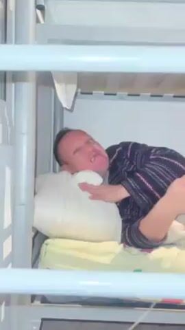 A man is lying in the bottom bunk of a bunk bed. He is wearing a patterned shirt and has his head on a pillow. He looks uncomfortable and makes a distressed sound. He then sees a phone recording him and points at the camera, scowling. The bunk bed is silver and white.