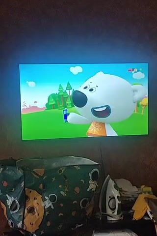 The video shows a television screen displaying an animated cartoon. In the first frame, a white bear character with a blue nose is holding a blue object to its mouth. The background is a vibrant green field with trees and a blue sky with white clouds. The second and third frames show a red fox character smiling and holding a notepad. The fourth frame shows the white bear character and a brown bear character hugging each other. In the foreground, partially visible, is a laundry hamper with an astronaut and moon print, and an iron.