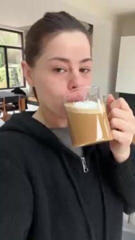 A woman with brown hair pulled back drinks from a large clear glass mug filled with a tan liquid, presumably coffee. She wears a gray shirt and a black zip-up hoodie. She smiles slightly after taking a sip. The background shows a bright, modern living space with a wooden dining table and chairs, a window looking out to greenery, and a staircase with a black railing.