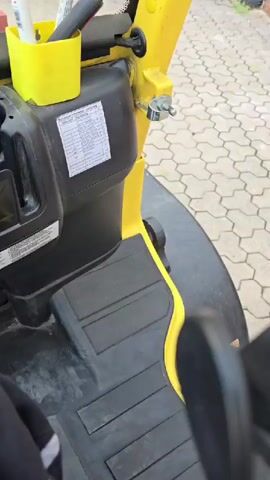The media displays a first-person perspective from the driver's seat of a forklift. The driver's legs are visible in the foreground, wearing black pants and shoes with green accents. The dashboard of the forklift is the primary focus, with various controls and a digital display. The display shows "Fuel" with a graphical indicator and the time "11:5" followed by a series of numbers that might represent an odometer reading, "204037". A green button is also visible on the dashboard.

The scene appears to be outdoors, with hints of a brick wall and paving stones visible through the forklift's windows. The forklift itself is yellow and black. A remote control with numerous buttons rests on a console. In the background, there are some boxes and a yellow bin. The overall impression is that of a working environment, possibly a warehouse or an industrial yard. The lighting suggests it is daytime, and the weather appears clear. There are no visible people other than the implied driver, and no strong emotional cues are conveyed. The time of day is explicitly indicated as 11:05. The location context of Haren (Ems), Germany, is provided externally.