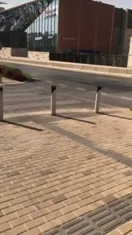 The video shows a sidewalk paved with light brown bricks, two short white and black posts, a crosswalk, and a road. In the background is a building with a store labeled "BURBERRY". A white SUV drives across the road.
