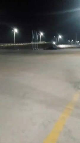 The video shows an outdoor scene at night. There are tall streetlights illuminating a concrete area, possibly a parking lot or an industrial space. In the background, there are flags on poles and some low-lying structures. A vehicle is visible in the distance. The camera appears to be moving, as indicated by the shifting perspective and motion blur. There are yellow painted lines on the concrete surface. The overall impression is one of an open, illuminated space at night.