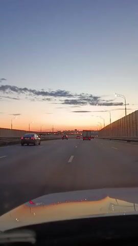 A vertical, first-person perspective video shot from inside a vehicle driving on a multi-lane highway. The sky is a gradient of pale blue to orange, indicating sunset or sunrise, with a few wispy clouds. Streetlights and a sound barrier line the right side of the highway. Several cars and a truck are visible on the road ahead. The interior of the vehicle, including the dashboard and windshield, is partially visible in the foreground. The overall lighting suggests dusk or dawn.