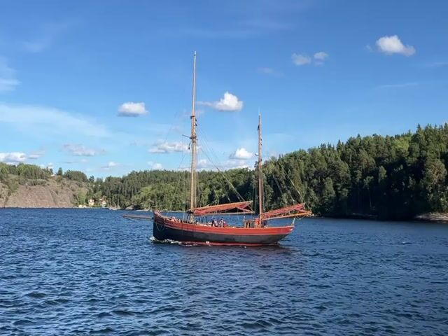 The video shows a two-masted sailboat with red and black hull, sailing on a blue body of water. The boat has several people on its deck. The background is a lush green forest that lines the shore of a hilly area. The sky is blue with scattered white clouds. The water is choppy, indicating the boat is in motion. The sun is bright, suggesting it is daytime. The overall scene depicts a pleasant day for sailing.
