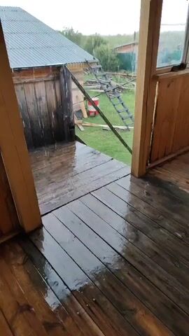 The video shows a wooden porch with wet floorboards due to rain. Beyond the porch, there is a wooden building with a metal roof and a ladder leaning against it. In the background, there is a green grassy area with some trees and other structures. The sky appears overcast. The rain is visible falling on the porch and the surrounding area. The footage is shaky, suggesting it was captured by someone holding the camera while moving.