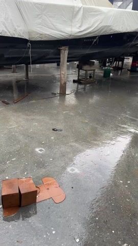 The video shows a wet concrete area, possibly a boatyard, with several boats resting on stands. Rain is falling and the ground is covered in puddles. One boat on the left is covered in a white tarp.  Two brown boxes and a piece of cardboard are in the foreground.  The lighting is overcast.