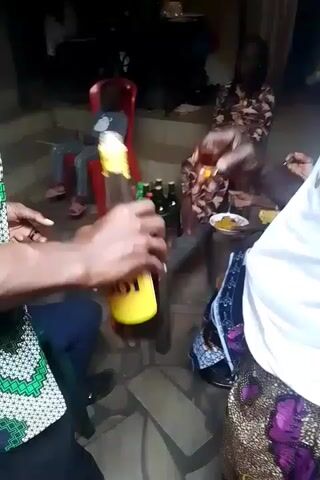 The video shows a gathering of people, primarily adults, in what appears to be an outdoor or semi-outdoor setting. There is a small child sitting on a red plastic chair. A table in the center holds an assortment of bottles, including what appear to be beers and wine, along with some glasses and food containers. People are eating from plates. Someone is seen opening a bottle. The environment seems relaxed and informal.