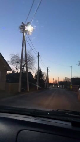 A car is driving down a narrow asphalt road in a rural area at dusk. The sky is a gradient of blue and orange, and the street is lined with wooden utility poles and houses. On top of some of the poles are large bird nests, some with birds visible. The car's headlights illuminate the road ahead. Music is playing in the background.