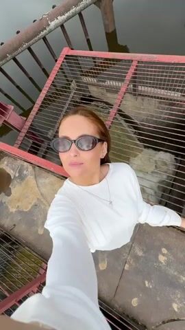 The video shows a woman with auburn hair and sunglasses standing on a concrete platform with a metal grate in front of her. She is wearing a white long-sleeved shirt and a necklace. Behind the grate, water is flowing. The woman turns her head to the side, and then looks back at the camera. The area appears to be a dam or some kind of water control structure.