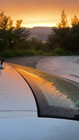 This is a vertical, first-person perspective video taken from the perspective of someone looking out of a car window at a sunset. The car is white and has water droplets on the hood and windshield. The hood of the car takes up the bottom third of the frame, with the windshield stretching up and to the right. The sunset is in the background, with hues of orange, yellow, and purple. Mountains and trees are silhouetted against the colorful sky. A dirt road or path is visible in the middle ground.