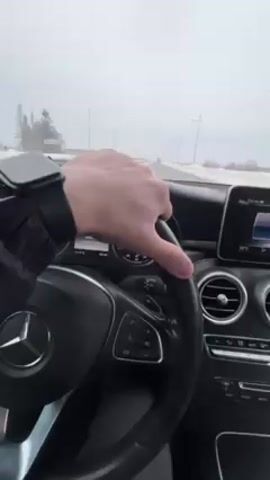 The video shows a person driving a Mercedes-Benz car on a snow-covered road. The driver's left hand is visible on the steering wheel. The car's dashboard and interior are also in view, including the speedometer and the infotainment screen. The weather appears to be overcast and snowy. The road is straight and flat, lined with snowy fields. There are utility poles on either side of the road.