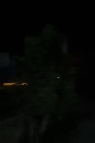 This video captures a nighttime scene from a moving vehicle. The camera is pointed forward and slightly to the left, showing a dark, unlit road. On the left side of the frame, there are dark trees and foliage. In the distance, on the right side of the road, there are several vehicles with their headlights on, illuminating the area. One vehicle, possibly a truck, has its brake lights illuminated red. Further ahead, there are some buildings or structures with bright lights emanating from them. The overall impression is that of a dimly lit rural or suburban road at night.