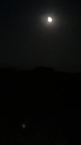 The video begins by showing a half-moon in a dark sky. The moon is the brightest object in the frame, and it appears to be setting or rising. The lower half of the frame is filled with darkness, suggesting a landscape or a body of water. As the video progresses, the moon moves slightly and becomes less bright. In the distance, faint lights appear along the horizon, suggesting the presence of a city or town. The lights are small and scattered, and they are not very bright. The video ends with the lights still visible on the horizon, and the sky is still dark.
