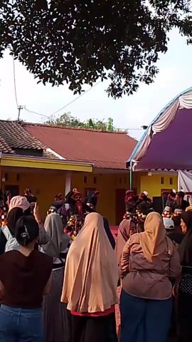 A group of people are gathered outdoors, watching a performance. A tent is set up to the right of the scene, and buildings are visible in the background. Several performers, dressed in traditional attire and masks, are on a raised platform or stage area, some of them dancing or gesturing. The audience members are facing the performers, some are holding up phones to record. The sky is visible above with trees in the foreground.