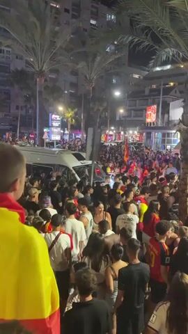 A large crowd of people are gathered on a street at night. Many of them are wearing red and yellow, the colors of the Spanish flag, and some are holding Spanish flags. There is a white van in the background, and palm trees and buildings are visible. The atmosphere appears to be celebratory, with people cheering and taking photos.