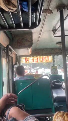 The video shows the interior of a bus. The camera is looking forward from a passenger's perspective. In the foreground, a person's hands are holding a smartphone, which is displaying colorful graphics. Behind them, other passengers are visible, including a man in a light blue shirt facing away from the camera, seated in a green seat. The bus appears to be in motion, as indicated by the blurred scenery outside the windows. There are various signs and luggage racks visible inside the bus. The overall environment suggests a public transportation setting, possibly in India, given the script visible on some of the signs. The lighting suggests daylight, and the weather appears to be clear or overcast.