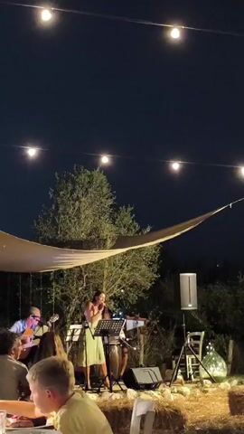 A woman sings into a microphone on a stage at night. She is wearing a light-colored dress. To her left, a man plays an acoustic guitar. There are other people visible in the background, and some audience members in the foreground. The stage is covered by a light-colored awning. String lights are visible above the scene. The ground is covered in straw bales. There are trees and bushes in the background. A speaker is set up to the right of the stage.