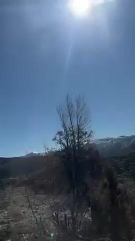 The video shows a rocky landscape under a bright, clear blue sky with the sun shining. A few barren trees are visible in the foreground, and a mountain range with some snow cover sits in the background. The camera appears to be moving, possibly from a car or other vehicle. The scene is bathed in strong sunlight, creating a high-contrast environment. There is music playing.