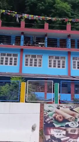 The video shows a side view of a moving vehicle, likely a bus, traveling along a road. Through the bus windows, a blue and pink building is visible, adorned with prayer flags and what appear to be air conditioning units on a wall. Signs on the building advertise "LE GRAND HOTEL FOOD KHALA" and "HOTEL NATRAJ". The environment is hilly with lush green trees.