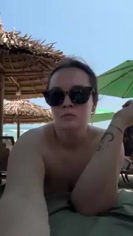 A woman is on a beach. She is wearing sunglasses and a watch. She has a tattoo on her left forearm. She is lying on a green towel under a straw umbrella. There is a green umbrella in the background. She is talking, looking directly at the camera.